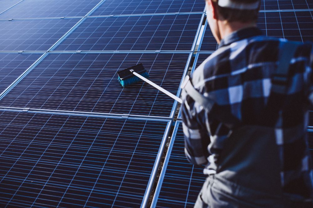 Mann reinigt mit einem langen Besen blaue Solarmodule